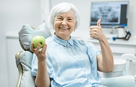 Older woman with dental implant retained dentures holding up a green apple and giving thumbs up