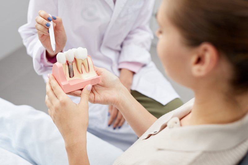 Woman in dental chair holding dental implant model as dentist points at different parts with a pen